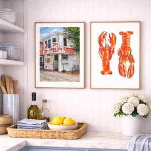 May include: Two framed art prints. One shows a white building with the text "Lobster Pot Steaks". The other features two watercolour illustrations of a red lobster. The art is displayed above a kitchen counter with a woven tray, lemons, and a vase of white flowers.