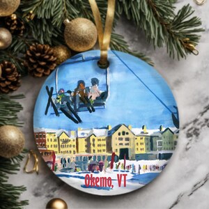 May include: A round, white, ceramic ornament with a painted scene of a ski lift with people riding it. The background shows a small town with buildings and a snowy landscape. The text "Okemo, VT" is printed on the bottom of the ornament.
