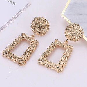 May include: Gold-toned dangle earrings with a textured, geometric design. The earrings feature a square-shaped drop with a smaller circle at the top.