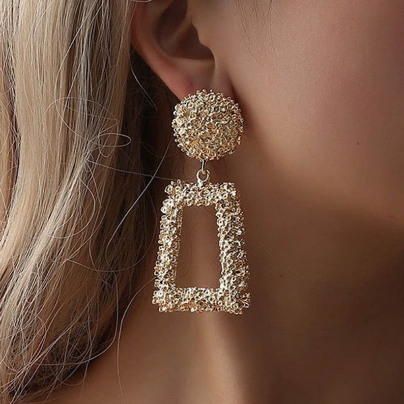 May include: Gold-tone statement earrings with a textured round top and a rectangular dangle. The earrings have a geometric design.