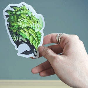 May include: A green dragon sticker with a watercolor effect. The dragon is facing to the right and has a detailed design with scales and spikes.