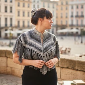 Puede incluir: Un chal de ganchillo gris y blanco con flecos. El chal tiene un cuello alto y un broche decorativo. La mujer de la imagen lleva un vestido negro y tiene el pelo recogido. El fondo es una plaza de ciudad europea.
