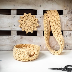 May include: Three crocheted items in a light yellow color: a round coaster, a bowl, and a long scarf. The items are displayed on a white surface with a wooden background.