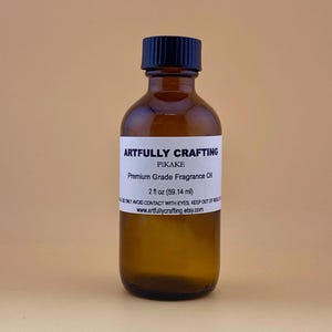 May include: A brown glass bottle with a black cap, labeled "ARTFULLY CRAFTING PIKAKE Premium Grade Fragrance Oil." The label indicates a 2 fl oz (59.14 ml) volume. The bottle is filled with a clear liquid.