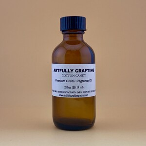 May include: A brown glass bottle with a black cap, labeled "Artfully Crafting Cotton Candy Premium Grade Fragrance Oil 2 fl oz (59.14 ml)".