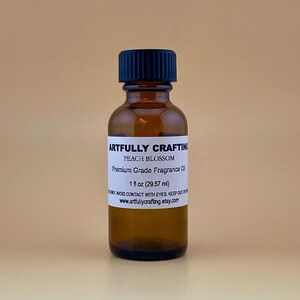 May include: A brown glass bottle with a black cap, labeled "ARTFULLY CRAFTING PEACH BLOSSOM Premium Grade Fragrance Oil 1 fl oz (29.57 ml)." The bottle is filled with a clear liquid.