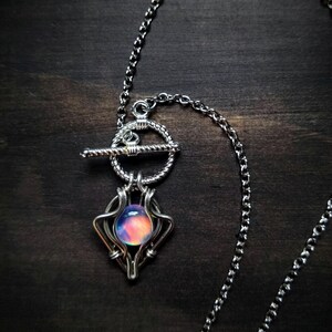 May include: Silver-toned necklace with a toggle clasp and a pendant featuring a colorful, iridescent orb. The pendant is encased in a wire-wrapped design. The chain and clasp are visible against a dark wooden surface.
