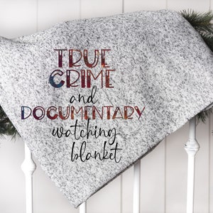 Puede incluir: Una manta gris y blanca moteada con el texto "TRUE CRIME and DOCUMENTARY watching blanket" en una fuente colorida. La manta está sobre una barandilla blanca.