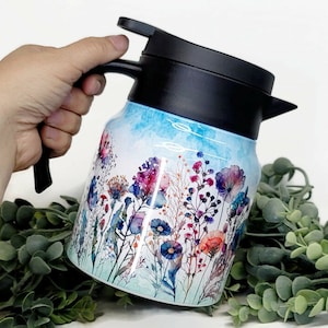 Puede incluir: Una jarra térmica de acero inoxidable azul y blanco con tapa y asa negras. La jarra presenta un diseño floral de acuarela con flores rosas, moradas y naranjas.