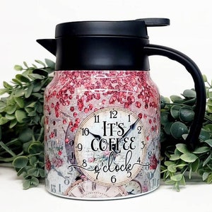 Op de afbeelding: Een roze en witte thermoskan voor koffie met een zwart deksel en handvat. De kan heeft een vintage wijzerplaat met de tekst "It's Coffee o'clock".
