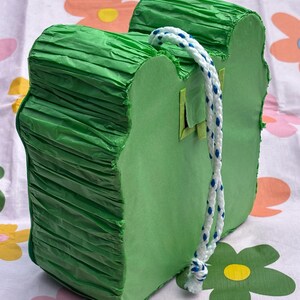 K&ouml;nnte beinhalten: Eine hellgr&uuml;ne Pi&ntilde;ata in Froschform mit flacher Vorder- und R&uuml;ckseite sowie Seiten aus gefaltetem gr&uuml;nem Papier. Ein wei&szlig;es, geflochtenes Seil mit blauen Sprenkeln ist oben zur Aufh&auml;ngung befestigt, und eine kleine quadratische Klappe ist sichtbar.