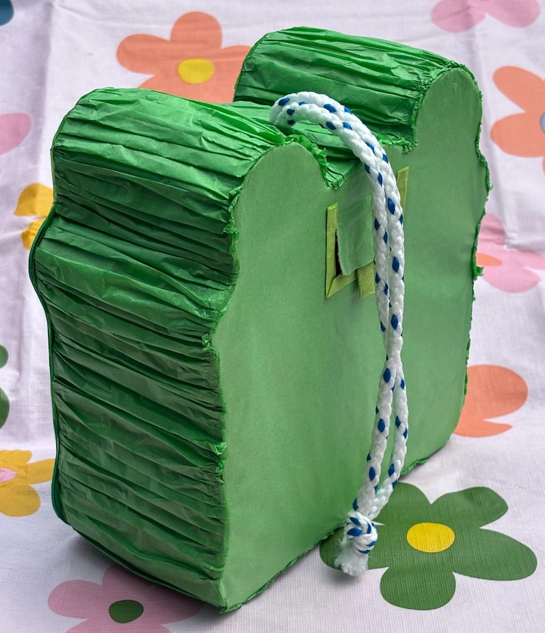 K&ouml;nnte beinhalten: Eine hellgr&uuml;ne Pi&ntilde;ata in Froschform mit flacher Vorder- und R&uuml;ckseite sowie Seiten aus gefaltetem gr&uuml;nem Papier. Ein wei&szlig;es, geflochtenes Seil mit blauen Sprenkeln ist oben zur Aufh&auml;ngung befestigt, und eine kleine quadratische Klappe ist sichtbar.