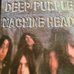 May include: Close-up of a vintage album cover for Deep Purple's "Machine Head." The album title is embossed in a silver, bubble-like font. The background features a blurred image of the band members.