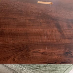 May include: A rectangular dark brown wooden board with a smooth, polished finish showing a prominent wood grain pattern. The board features two pre-cut keyhole slots on the back surface, designed for wall mounting, and a thin vertical crack.