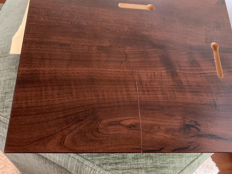 May include: A rectangular dark brown wooden board with a smooth, polished finish showing a prominent wood grain pattern. The board features two pre-cut keyhole slots on the back surface, designed for wall mounting, and a thin vertical crack.