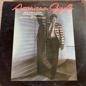 May include: A black vinyl record album cover with a photo of a man in a striped suit standing in a room with a large window. The album cover is for the soundtrack to the film "American Gigolo". The text on the cover reads "American Gigolo", "Original Soundtrack Recording", "Music Composed by Giorgio Moroder", "featuring", "Blondie / "Call Me"", and "Cheryl Barnes / "Love & Passion"".