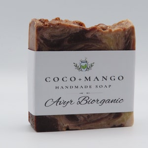May include: A bar of handmade soap with a white label. The soap is a mix of brown and tan colors with a textured top. The label reads "COCO + MANGO HANDMADE SOAP" and "Avyr Biorganic".