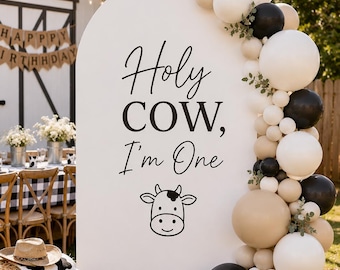 May include: A white arched backdrop featuring the black text Holy COW, I'm One and a cow illustration. A balloon garland in white, black, and beige wraps around the right side. The display is set outdoors on grass with a hay bale and boots nearby.