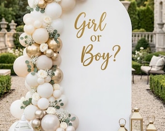 May include: A white arched backdrop featuring the gold text Girl or Boy? decorated with a balloon garland in shades of cream, white, and metallic gold, accented with small white flowers and green leaves, plus four metal lanterns on the ground.