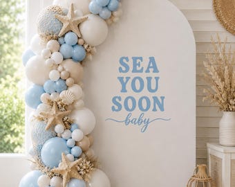 May include: A white arched backdrop featuring the blue text SEA YOU SOON baby, decorated with a balloon garland in shades of light blue, white, and beige, accented with dried grasses, a white seashell, and three large tan starfish.