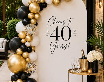 May include: A white arched backdrop featuring the black text Cheers to 40 Years! decorated with a balloon garland in black, white, gold, and clear confetti-filled balloons. A gold bar cart with champagne glasses and a bottle sits to the right.