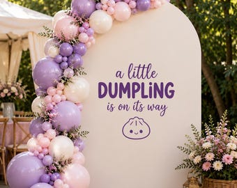 May include: A white arched backdrop featuring a balloon garland in shades of lavender, light pink, and white. The text reads "a little DUMPLING is on its way" in purple, accompanied by a small, smiling dumpling graphic centered on the panel.