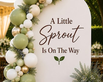 May include: A white arched backdrop featuring the brown text A Little Sprout Is On The Way with a green leaf icon. A balloon garland in shades of sage green, white, and metallic gold is attached to the left side, accented with green fern foliage.