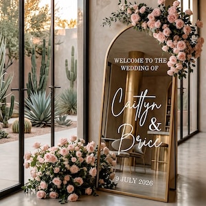 Puede incluir: Un espejo de pie arqueado con marco dorado que presenta texto en blanco que dice Welcome to the wedding of Caitlyn & Brice 9 July 2026. El espejo está decorado con racimos de rosas de color rosa claro en la parte superior derecha y la base.