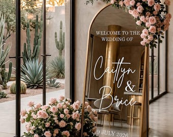 May include: An arched floor mirror with a gold frame featuring white text that reads Welcome to the wedding of Caitlyn & Brice 9 July 2026. The mirror is decorated with clusters of light pink roses at the top right and base, reflecting a room.