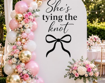 May include: A white arched backdrop featuring the black text She's tying the knot above a black bow graphic. A balloon garland in shades of pink, white, and metallic gold is accented with pink roses and greenery. A floral arrangement sits nearby.
