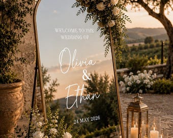 May include: An arched gold-framed mirror wedding sign featuring white text that reads WELCOME TO THE WEDDING OF Olivia & Ethan 24 MAY 2026. The mirror is decorated with white roses and greenery at the top and base, reflecting a sunset landscape.