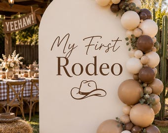 May include: A large, arched, cream-colored backdrop featuring the brown text 'My First Rodeo' and a cowboy hat graphic. A balloon garland in shades of cream, tan, and dark brown is attached to the right side, accented with small green leaves.