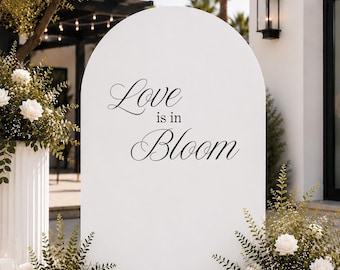 May include: A large white arched sign featuring the black text Love is in Bloom. The sign is surrounded by arrangements of white roses, green foliage, and clear glass cylinder candle holders containing white pillar candles on the ground.