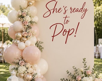 May include: An arched beige backdrop featuring the red text She's ready to Pop! decorated with a balloon garland in cream, blush, and metallic gold, accented by white and pink floral arrangements, a white lantern, and a woven basket.