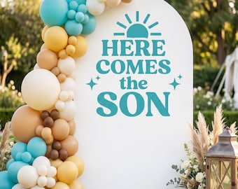 May include: A white arched backdrop featuring the teal text "HERE COMES THE SON" with a sun icon and star accents. A balloon garland in shades of teal, tan, cream, and brown is attached to the left side of the arch, creating a decorative display.