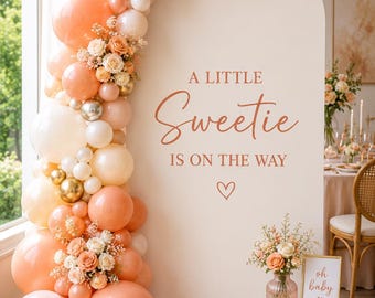 May include: A large white arched backdrop featuring the text A LITTLE Sweetie IS ON THE WAY with a heart icon. The arch is decorated with a balloon garland in shades of peach, cream, and gold, accented with small white and peach floral arrangements.