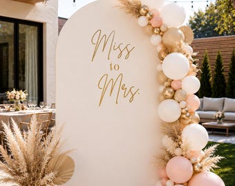 May include: A white arched backdrop featuring the gold text "Miss to Mrs." decorated with a balloon garland in white, blush pink, and metallic gold, accented with dried pampas grass and palm leaves, alongside a ceramic vase with pampas grass.