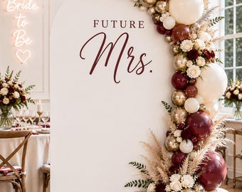 May include: A white arched backdrop featuring the text FUTURE Mrs. in dark red lettering. The arch is decorated with a balloon garland in shades of burgundy, cream, and metallic gold, accented with dried pampas grass and white floral blooms.