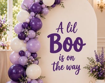 May include: A large white arched backdrop featuring the purple text A lil Boo is on the way. A balloon garland in shades of dark purple, light purple, and white is attached to the left side, accented with small sprigs of purple flowers and greenery.