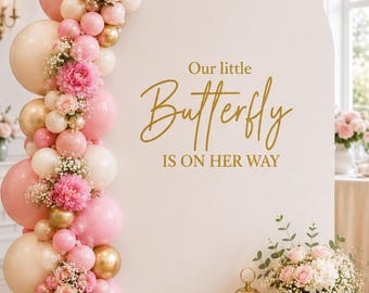 May include: A white arched backdrop featuring a balloon garland in shades of pink, cream, and gold, accented with pink flowers. The text "Our little Butterfly IS ON HER WAY" is printed in gold. A gold lantern with a candle and a vase of flowers sit below.