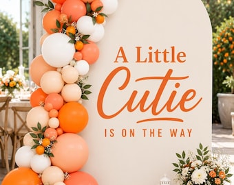 May include: A large arched backdrop in cream featuring the orange text A Little Cutie IS ON THE WAY. A balloon garland in shades of orange, peach, and white is attached to the left side, accented with small green leaves and tiny orange fruits.