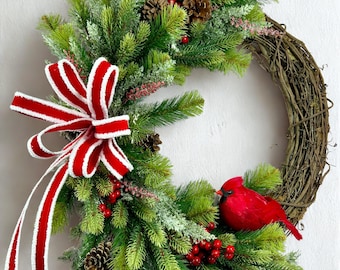 May include: A Christmas wreath with a natural grapevine base, adorned with green pine needles, red berries, and pinecones. A red and white striped bow and a red cardinal add festive touches. This holiday decoration is perfect for the front door.