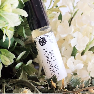 May include: A perfume oil sample vial with a black cap and a white label that reads "Salt Air + Honey Dew". The vial is surrounded by white and green flowers. The text "Perfume Sample Vials Set of (5)" is at the bottom.