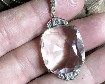 May include: A vintage pendant necklace featuring a large, clear, faceted gemstone. The pendant is rectangular with rounded corners, set in a silver-toned metal frame with small accent stones. The necklace has a delicate chain.