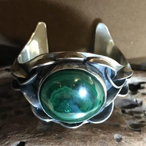 May include: A silver cuff bracelet featuring a large, round, polished malachite stone. The bracelet has a floral design around the stone and flared edges. The malachite stone displays a rich green color with darker green patterns.