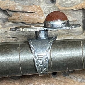 May include: A silver ring with a reddish-brown stone set on top. The ring has a unique design with a wide, decorative band and a detailed setting for the stone. The ring is displayed on a metal ring sizer.