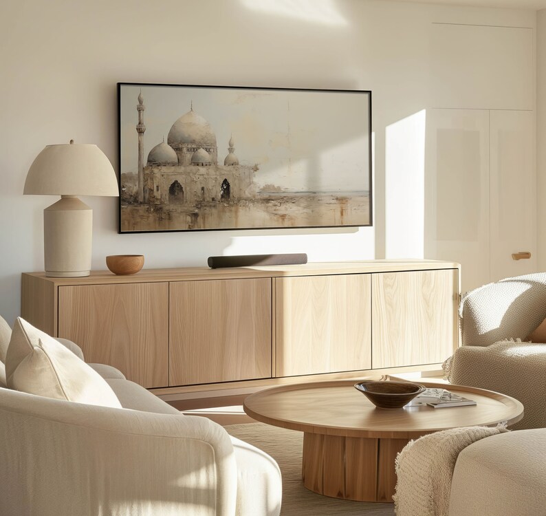 May include: A living room with a light wood cabinet, a white sofa, a coffee table, and a lamp. A flat screen TV is mounted on the wall above the cabinet. The TV is displaying a painting of a mosque.