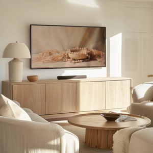 May include: A light wood cabinet with three drawers and a flat screen TV above it. A white lamp with a white lampshade is on the left side of the cabinet. A light wood coffee table with a bowl on top is in front of the cabinet. A white sofa is in the foreground.