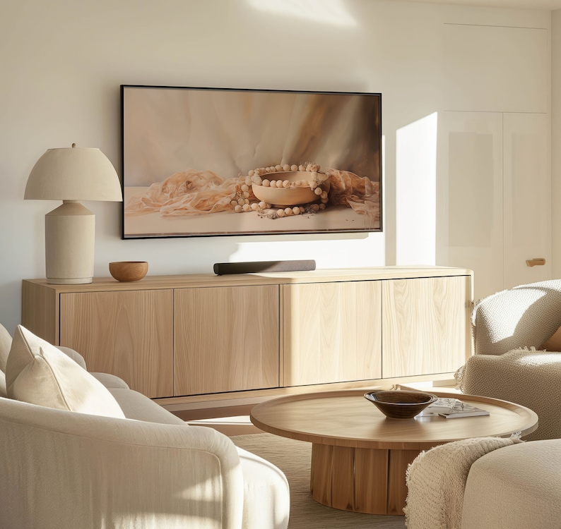 May include: A light wood cabinet with three drawers and a flat screen TV above it. A white lamp with a white lampshade is on the left side of the cabinet. A light wood coffee table with a bowl on top is in front of the cabinet. A white sofa is in the foreground.