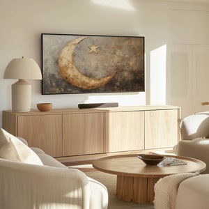 May include: A living room featuring a light wood cabinet, a coffee table, and a sofa. The cabinet has a large flat screen TV on top of it. The TV is displaying a painting of a crescent moon and a star. The coffee table is round and has a wooden top. The sofa is light coloured and has a white throw pillow on it.